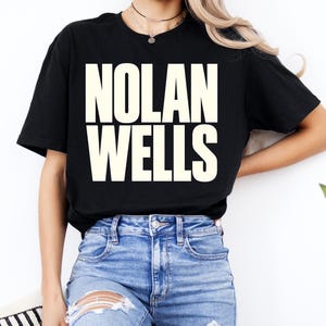 May include: A black short-sleeve t-shirt featuring the text NOLAN WELLS printed in large, bold, cream-colored capital letters across the chest. The shirt is paired with blue denim jeans that have distressed rips on the front thigh areas.