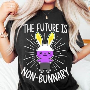 Pode incluir: Uma t-shirt preta com um gráfico de um coelho de desenho animado nas cores amarelo, branco, roxo e preto. O texto acima e abaixo do coelho diz THE FUTURE IS NON-BUNNARY, rodeado por linhas brancas radiantes num fundo escuro.