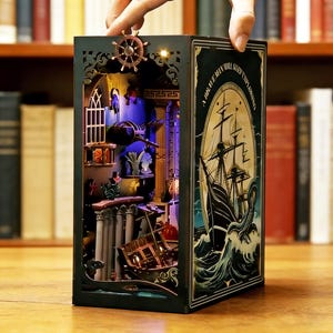 May include: A decorative, illuminated book nook with a nautical theme. The interior features a detailed miniature scene with columns, a ship, and a shipwreck. The exterior has a ship illustration and the text "A Brave Man Will Keep Exploring."