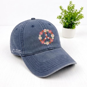 May include: A denim blue baseball cap with a floral peace sign embroidered on the front. The peace sign is made of pink, white, and yellow flowers. The text "No Kings In America" is on the side.