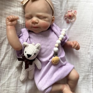 Pu&ograve; includere: Una bambola neonata realistica con gli occhi chiusi, che indossa una tutina a maniche corte color viola chiaro e una fascia beige con fiocco. La bambola tiene in mano un piccolo orsetto all'uncinetto color crema con un nastro marrone scuro.
