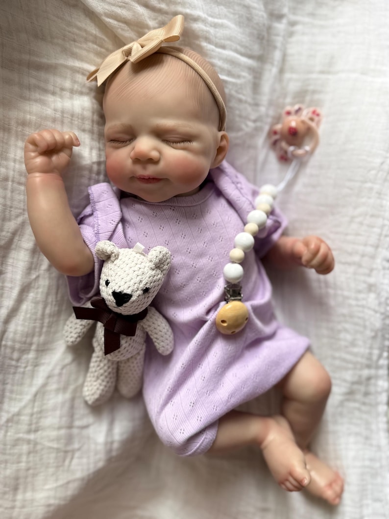 Pu&ograve; includere: Una bambola neonata realistica con gli occhi chiusi, che indossa una tutina a maniche corte color viola chiaro e una fascia beige con fiocco. La bambola tiene in mano un piccolo orsetto all'uncinetto color crema con un nastro marrone scuro.