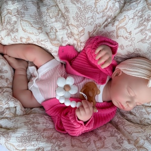 May include: A baby doll wearing a pink knitted sweater and striped shorts, lying on a patterned blanket. The doll has a white headband and is holding a wooden teether. The teether is shaped like a flower and a bear.