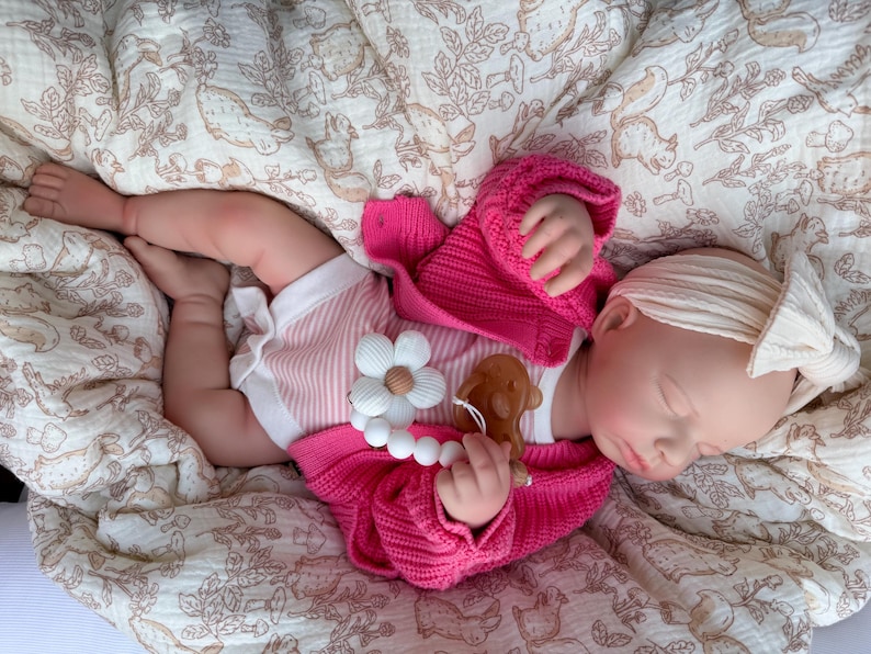 May include: A baby doll wearing a pink knitted sweater and striped shorts, lying on a patterned blanket. The doll has a white headband and is holding a wooden teether. The teether is shaped like a flower and a bear.