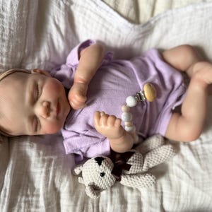 Pu&ograve; includere: Una bambola neonato realistica con gli occhi chiusi, che indossa un body a maniche corte color viola chiaro e una fascia beige con un fiocco. La bambola ha un portaciuccio con perline bianche e beige e tiene in mano un piccolo orsetto all'uncinetto.