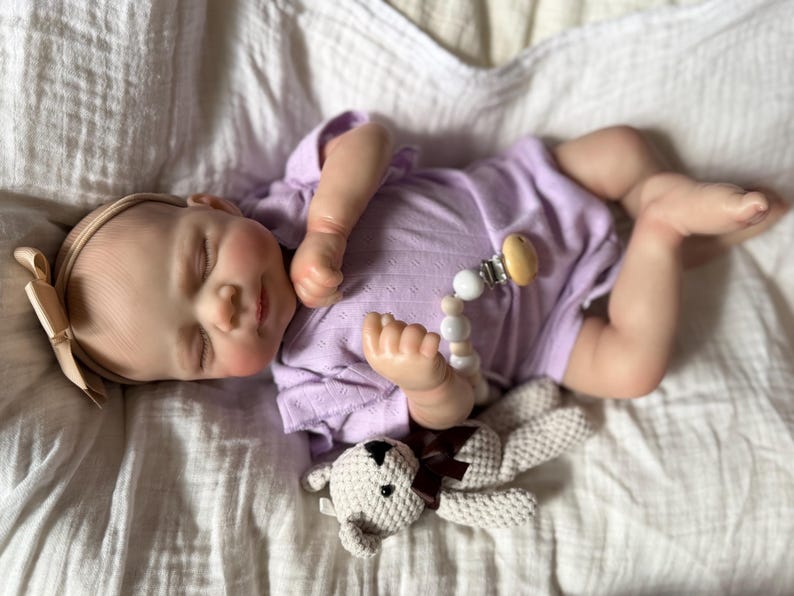 Pu&ograve; includere: Una bambola neonato realistica con gli occhi chiusi, che indossa un body a maniche corte color viola chiaro e una fascia beige con un fiocco. La bambola ha un portaciuccio con perline bianche e beige e tiene in mano un piccolo orsetto all'uncinetto.