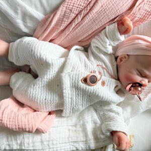 May include: A realistic baby doll with light skin, wearing a white textured long-sleeve top and matching pants with subtle grey dots. The doll has a pink headband with a bow, a rose gold pacifier, and a rose gold pacifier clip attached to the top.