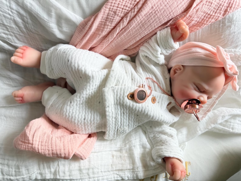 May include: A realistic baby doll with light skin, wearing a white textured long-sleeve top and matching pants with subtle grey dots. The doll has a pink headband with a bow, a rose gold pacifier, and a rose gold pacifier clip attached to the top.
