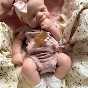 Może przedstawiać: Realistyczna lalka niemowlę z zamkniętymi oczami, ubrana w prążkowane body z kr&oacute;tkim rękawem w kolorze przybrudzonego r&oacute;żu, pasującą r&oacute;żową opaskę z kokardą oraz białe skarpetki z falbankami. Smoczek w kolorze beżowym jest przypięty do body.