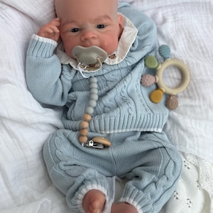 K&ouml;nnte beinhalten: Eine realistische Babypuppe mit heller Haut tr&auml;gt einen hellblauen Zopfstrickpullover und eine passende Hose mit wei&szlig;em Saum. Die Puppe hat einen hellgr&uuml;nen Schnuller an einem Silikonperlenclip und h&auml;lt einen runden, mehrfarbigen Bei&szlig;ring.