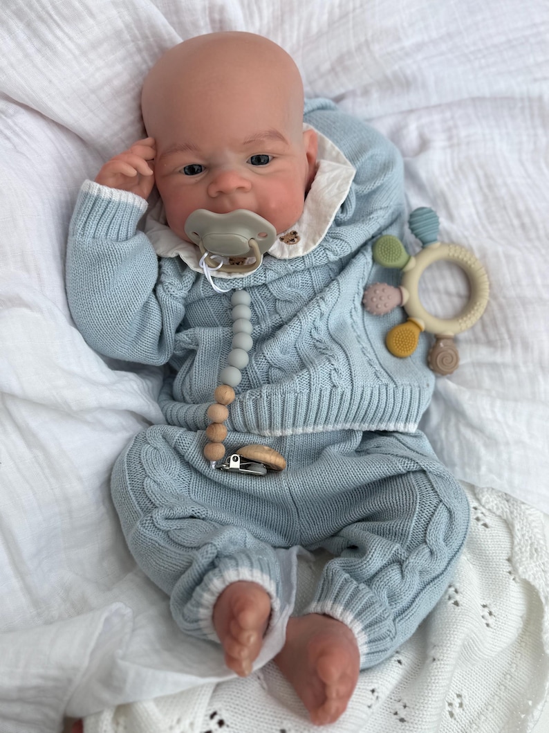 K&ouml;nnte beinhalten: Eine realistische Babypuppe mit heller Haut tr&auml;gt einen hellblauen Zopfstrickpullover und eine passende Hose mit wei&szlig;em Saum. Die Puppe hat einen hellgr&uuml;nen Schnuller an einem Silikonperlenclip und h&auml;lt einen runden, mehrfarbigen Bei&szlig;ring.