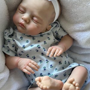 May include: A realistic reborn doll with light skin, closed eyes, and painted hair, wearing a white short-sleeved onesie with a small blue and green floral pattern and a white headband featuring a fabric flower, resting on a soft white fabric.
