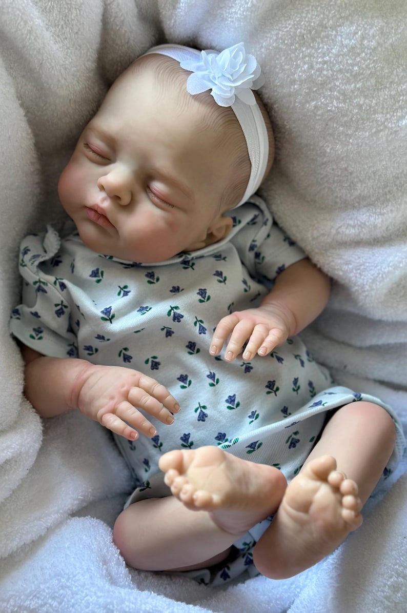 May include: A realistic reborn doll with light skin, closed eyes, and painted hair, wearing a white short-sleeved onesie with a small blue and green floral pattern and a white headband featuring a fabric flower, resting on a soft white fabric.