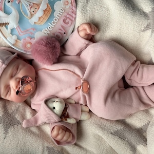 Pu&ograve; includere: Una bambola reborn realistica che indossa una tutina a maniche lunghe a coste rosa chiaro e un berretto abbinato. La bambola ha un ciuccio in oro rosa, una clip per ciuccio in oro rosa e un piccolo peluche. &Egrave; visibile un cartello circolare con l'illustrazione di una cicogna e il testo Welcome Baby Girl.