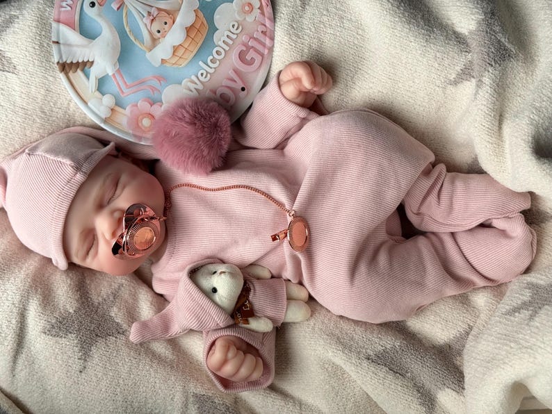Pu&ograve; includere: Una bambola reborn realistica che indossa una tutina a maniche lunghe a coste rosa chiaro e un berretto abbinato. La bambola ha un ciuccio in oro rosa, una clip per ciuccio in oro rosa e un piccolo peluche. &Egrave; visibile un cartello circolare con l'illustrazione di una cicogna e il testo Welcome Baby Girl.