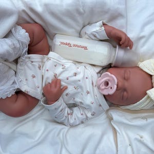 May include: A lifelike baby doll dressed in a white floral onesie, socks, and a cream bow headband. A pink dummy is in its mouth, and a bottle of formula rests beside its head. The doll is lying on a white blanket.