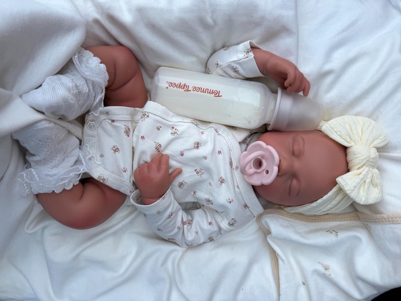 May include: A lifelike baby doll dressed in a white floral onesie, socks, and a cream bow headband. A pink dummy is in its mouth, and a bottle of formula rests beside its head. The doll is lying on a white blanket.