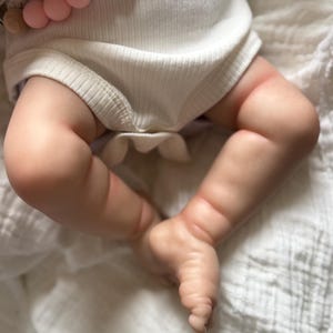May include: A realistic doll with light skin tone wearing a white ribbed bodysuit. A string of round, light pink silicone beads is draped across the doll's torso. The doll's legs are bent at the knees, showing detailed skin texture and creases.