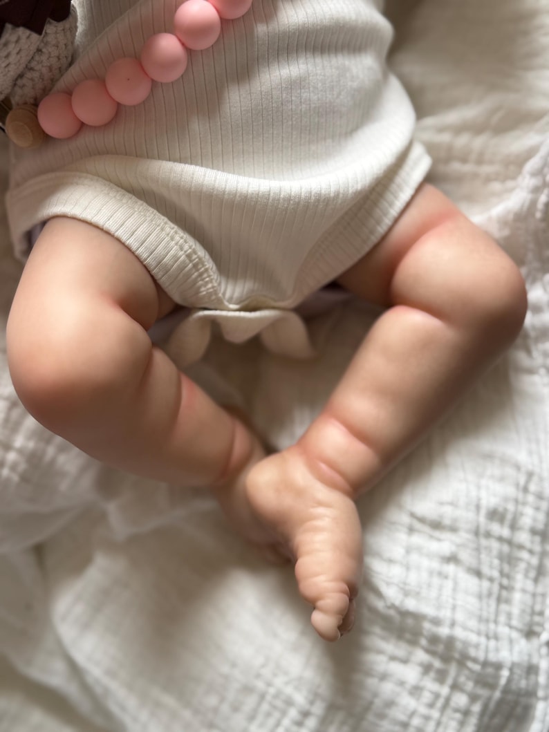 May include: A realistic doll with light skin tone wearing a white ribbed bodysuit. A string of round, light pink silicone beads is draped across the doll's torso. The doll's legs are bent at the knees, showing detailed skin texture and creases.