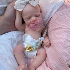 May include: A realistic newborn doll with light skin, closed eyes, and a sleeping expression. The doll wears a white diaper, a beige fabric bow headband, and a pink pacifier attached to a beige beaded pacifier clip with a gold metal clasp.