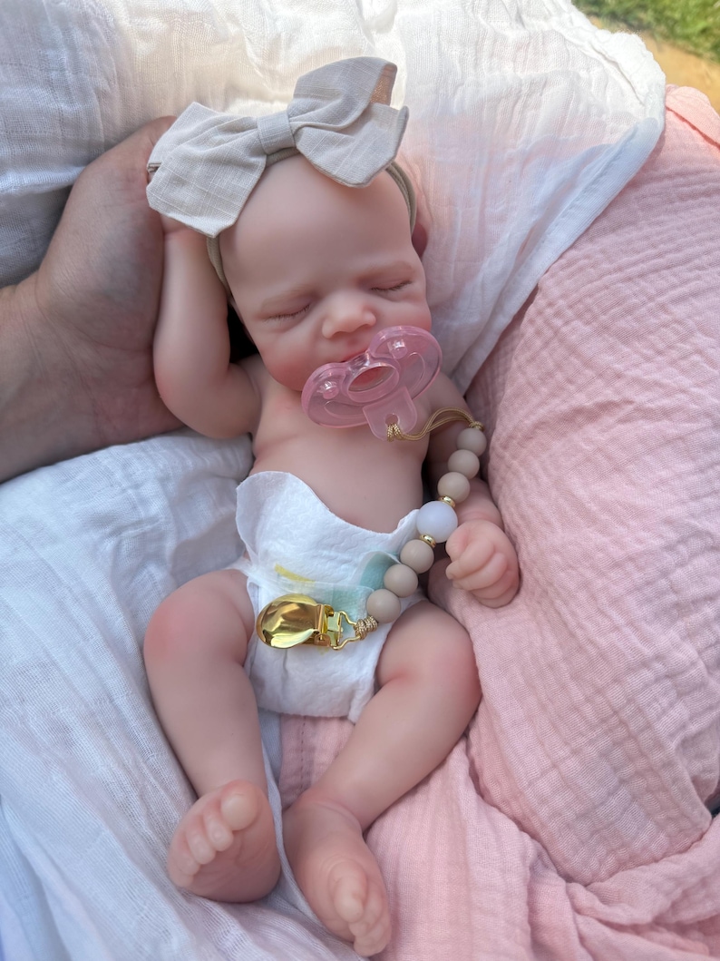 May include: A realistic newborn doll with light skin, closed eyes, and a sleeping expression. The doll wears a white diaper, a beige fabric bow headband, and a pink pacifier attached to a beige beaded pacifier clip with a gold metal clasp.