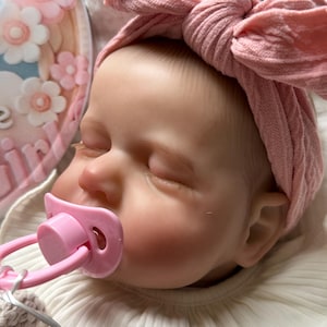 May include: A realistic baby doll with closed eyes, wearing a dusty pink cable-knit headband with a large bow. The doll has a light pink pacifier in its mouth and is dressed in a white ribbed shirt with a ruffled collar. A floral plaque is visible.