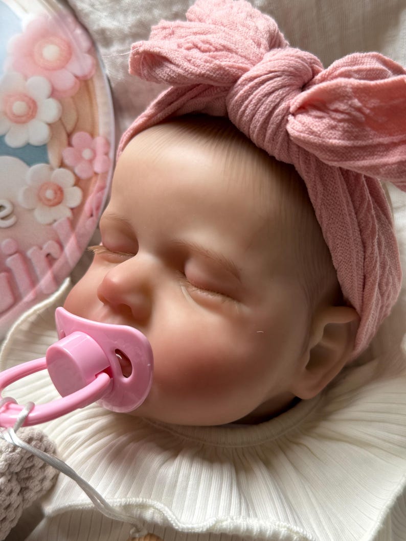 May include: A realistic baby doll with closed eyes, wearing a dusty pink cable-knit headband with a large bow. The doll has a light pink pacifier in its mouth and is dressed in a white ribbed shirt with a ruffled collar. A floral plaque is visible.