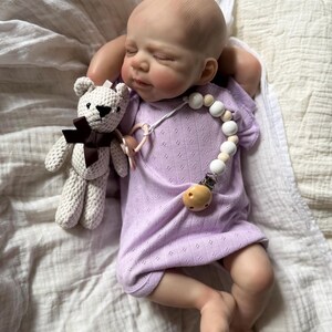 Pu&ograve; includere: Una bambola neonato realistica con testa calva e occhi chiusi, che indossa una tutina a maniche corte color viola chiaro con un sottile motivo pointelle. La bambola tiene in mano un piccolo orsacchiotto all'uncinetto color crema con un fiocco marrone.