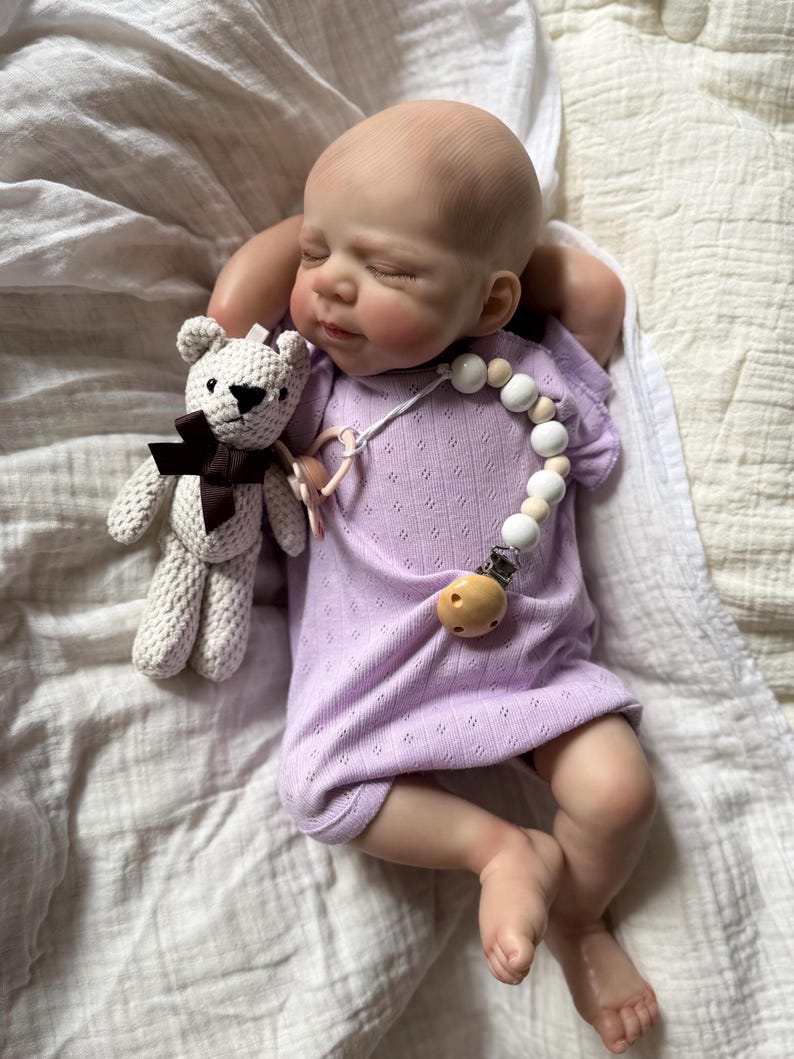 Pu&ograve; includere: Una bambola neonato realistica con testa calva e occhi chiusi, che indossa una tutina a maniche corte color viola chiaro con un sottile motivo pointelle. La bambola tiene in mano un piccolo orsacchiotto all'uncinetto color crema con un fiocco marrone.