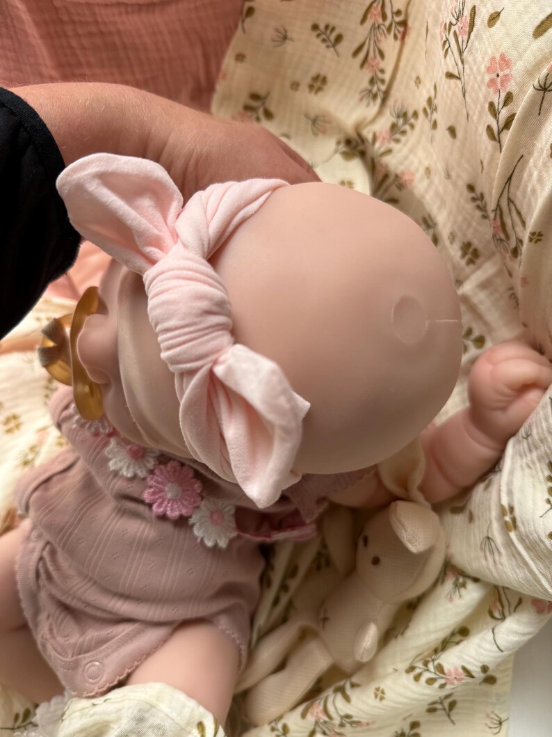 Może przedstawiać: Lalka o gładkiej, jasnej głowie nosi miękką, jasnor&oacute;żową opaskę z węzłem. Lalka ubrana jest w brudnor&oacute;żowe body z rzędem małych białych i r&oacute;żowych kwiatowych aplikacji przy dekolcie oraz złotym smoczkiem.