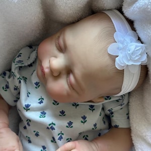 May include: A realistic baby doll with light skin, closed eyes, and long eyelashes, wearing a white headband with a fabric flower. The doll is dressed in a white short-sleeved shirt featuring a repeating pattern of small blue and green flowers.