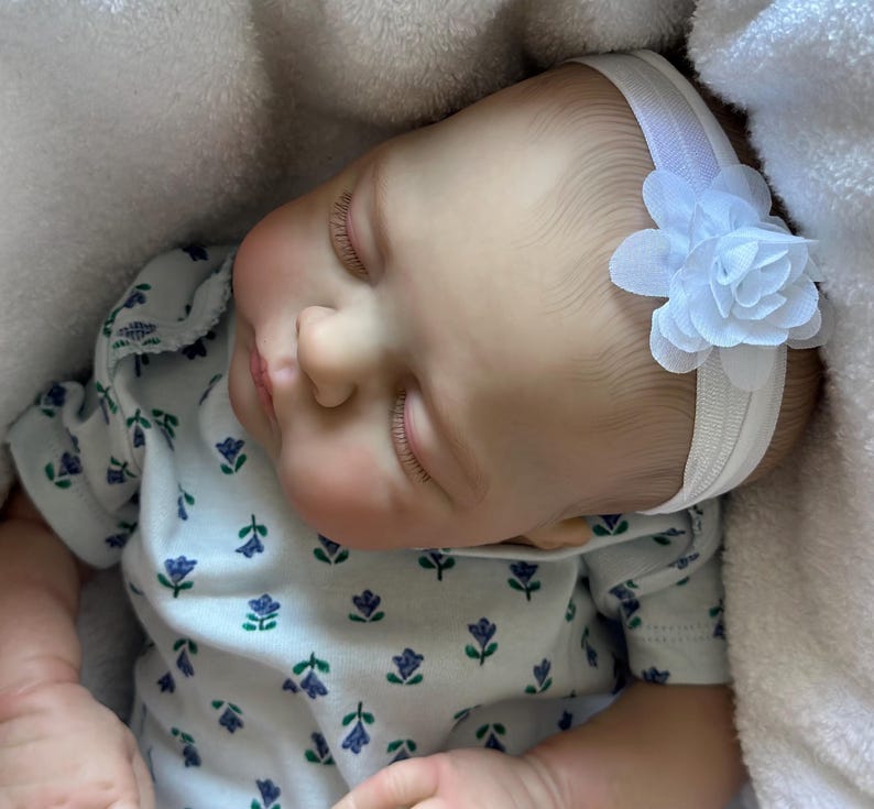 May include: A realistic baby doll with light skin, closed eyes, and long eyelashes, wearing a white headband with a fabric flower. The doll is dressed in a white short-sleeved shirt featuring a repeating pattern of small blue and green flowers.