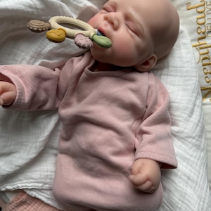 Pu&ograve; includere: Una bambola realistica dalla carnagione chiara e liscia, che indossa una tutina a maniche lunghe a coste color rosa polvere. La bambola ha gli occhi chiusi e tiene in mano un anello da dentizione circolare in silicone multicolore.