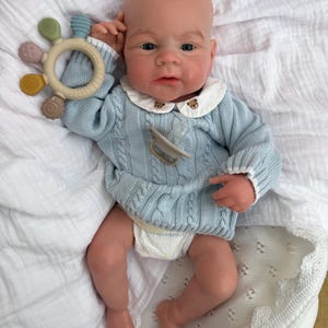 K&ouml;nnte beinhalten: Eine realistische Babypuppe mit heller Haut, bekleidet mit einem hellblauen Zopfmuster-Pullover mit wei&szlig;em Kragen, der kleine B&auml;renstickereien aufweist, und einer Windel. Die Puppe hat einen beigen Schnuller und h&auml;lt einen runden Silikon-Bei&szlig;ring.