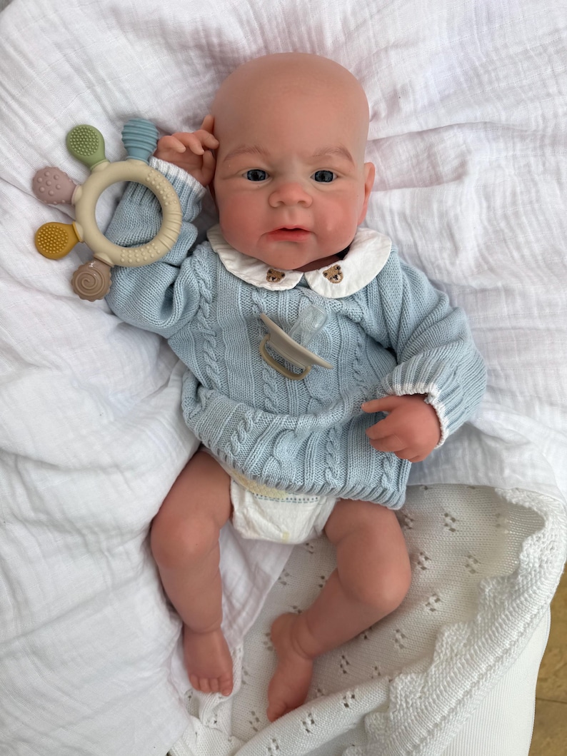 K&ouml;nnte beinhalten: Eine realistische Babypuppe mit heller Haut, bekleidet mit einem hellblauen Zopfmuster-Pullover mit wei&szlig;em Kragen, der kleine B&auml;renstickereien aufweist, und einer Windel. Die Puppe hat einen beigen Schnuller und h&auml;lt einen runden Silikon-Bei&szlig;ring.