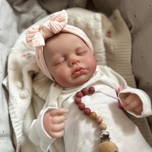 May include: A realistic baby doll with closed eyes, wearing a pale pink cable-knit headband and a plain white long-sleeved onesie. A pacifier clip with round, alternating terracotta and wooden beads is attached to the front of the onesie.