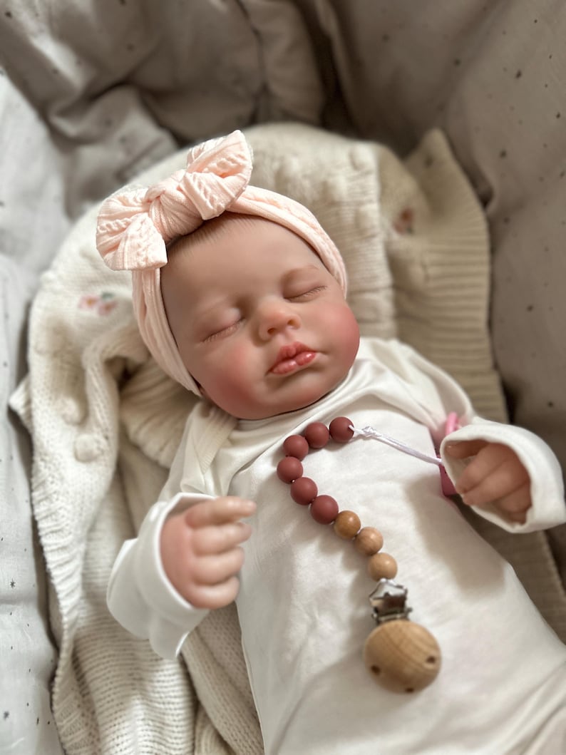May include: A realistic baby doll with closed eyes, wearing a pale pink cable-knit headband and a plain white long-sleeved onesie. A pacifier clip with round, alternating terracotta and wooden beads is attached to the front of the onesie.
