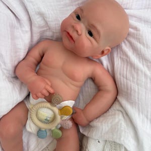 K&ouml;nnte beinhalten: Eine realistische Neugeborenen-Puppe mit heller Haut, kahlem Kopf und dunklen Augen, die eine wei&szlig;e Windel tr&auml;gt. Die Puppe h&auml;lt einen kreisf&ouml;rmigen, mehrfarbigen Silikon-Bei&szlig;ring mit kleinen, texturierten Noppen in Braun, Gelb, Rosa und Blau.