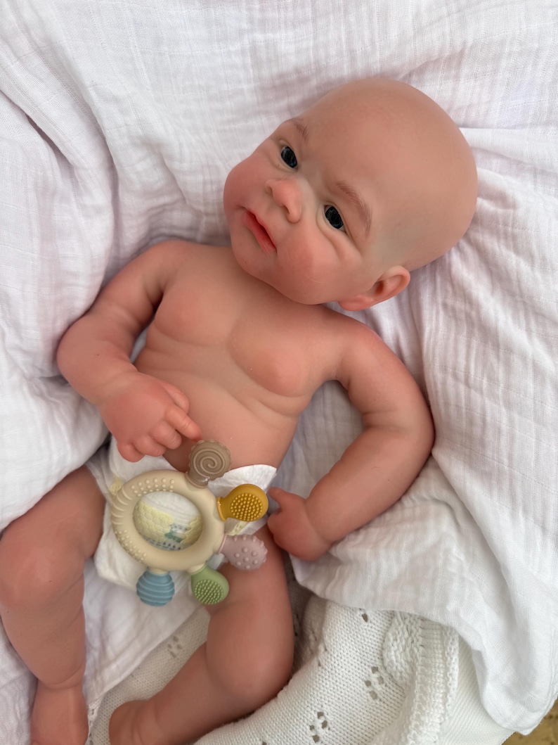 K&ouml;nnte beinhalten: Eine realistische Neugeborenen-Puppe mit heller Haut, kahlem Kopf und dunklen Augen, die eine wei&szlig;e Windel tr&auml;gt. Die Puppe h&auml;lt einen kreisf&ouml;rmigen, mehrfarbigen Silikon-Bei&szlig;ring mit kleinen, texturierten Noppen in Braun, Gelb, Rosa und Blau.