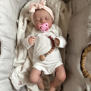May include: A realistic baby doll with closed eyes, wearing a white long-sleeved bodysuit and a pale pink knit headband with a bow. The doll has a pink pacifier attached to a beaded pacifier clip featuring brown and tan beads and a wooden clip.