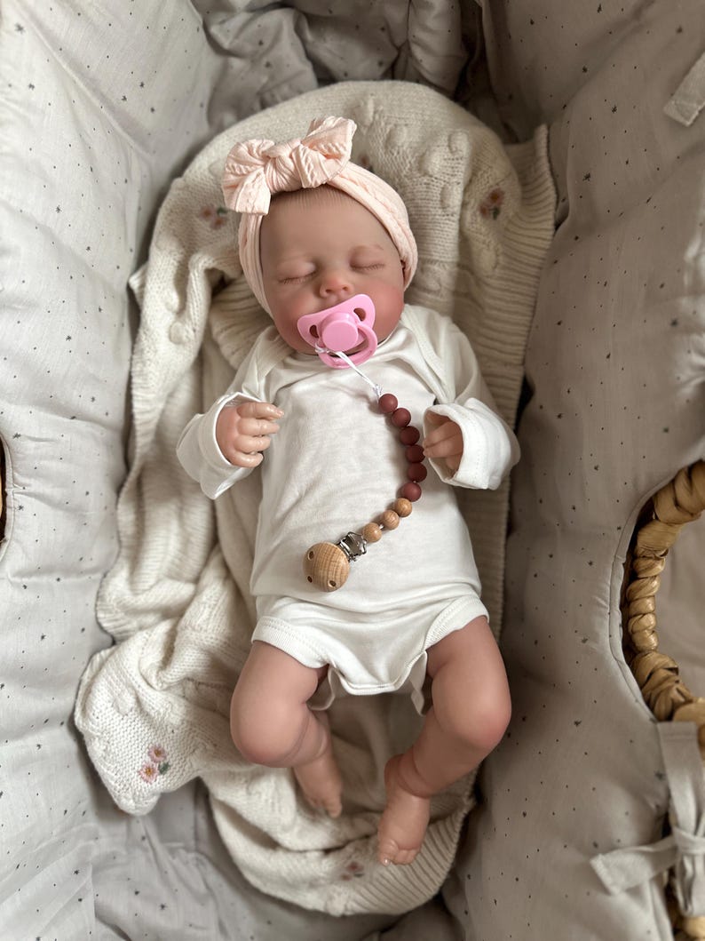 May include: A realistic baby doll with closed eyes, wearing a white long-sleeved bodysuit and a pale pink knit headband with a bow. The doll has a pink pacifier attached to a beaded pacifier clip featuring brown and tan beads and a wooden clip.