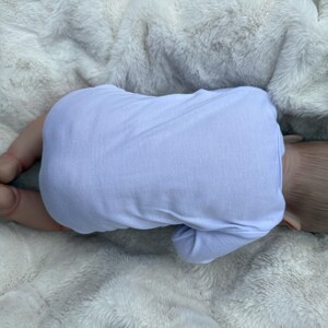 May include: A realistic baby doll with light brown hair, positioned lying on its stomach. The doll is wearing a plain, long-sleeved, light lavender-colored onesie that covers its torso and arms, while its bare legs and feet are visible.