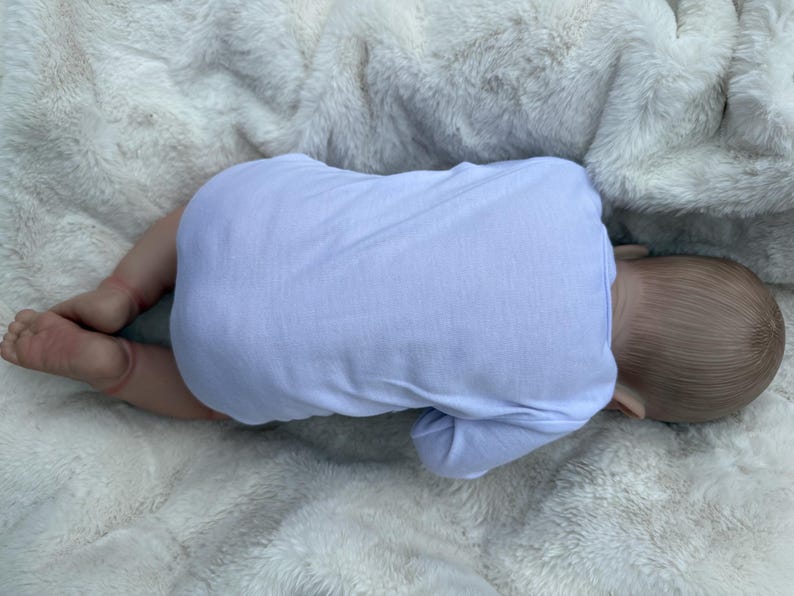 May include: A realistic baby doll with light brown hair, positioned lying on its stomach. The doll is wearing a plain, long-sleeved, light lavender-colored onesie that covers its torso and arms, while its bare legs and feet are visible.