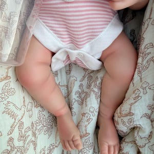 May include: A baby wearing a pink and white striped onesie with white trim. The baby is lying on a cream-colored blanket with a brown floral and animal print. A white flower-shaped pacifier holder is visible.