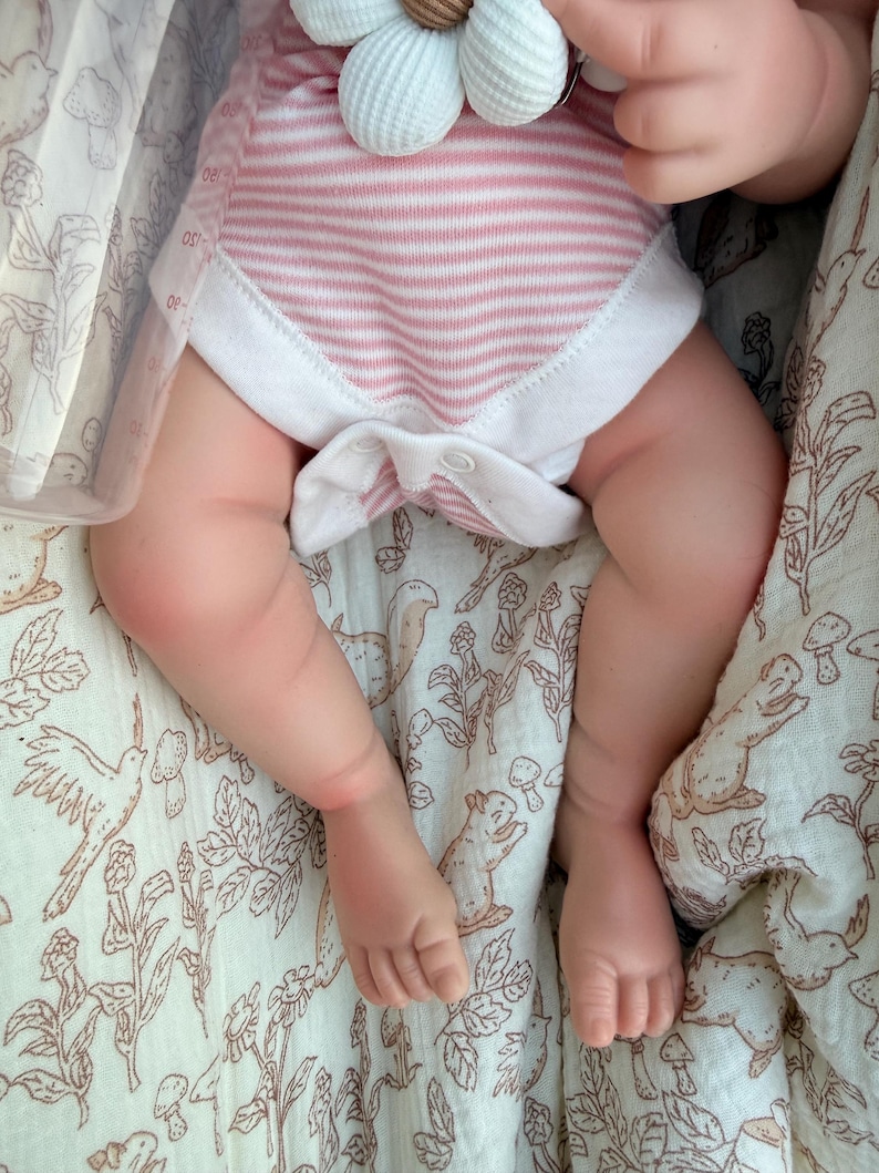 May include: A baby wearing a pink and white striped onesie with white trim. The baby is lying on a cream-colored blanket with a brown floral and animal print. A white flower-shaped pacifier holder is visible.
