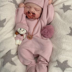 Pu&ograve; includere: Una bambola neonato addormentata che indossa una tutina a coste rosa polvere a maniche lunghe con berretto abbinato. La bambola ha un ciuccio color oro rosa con catenella e tiene in mano un piccolo orsacchiotto bianco con berretto e sciarpa.