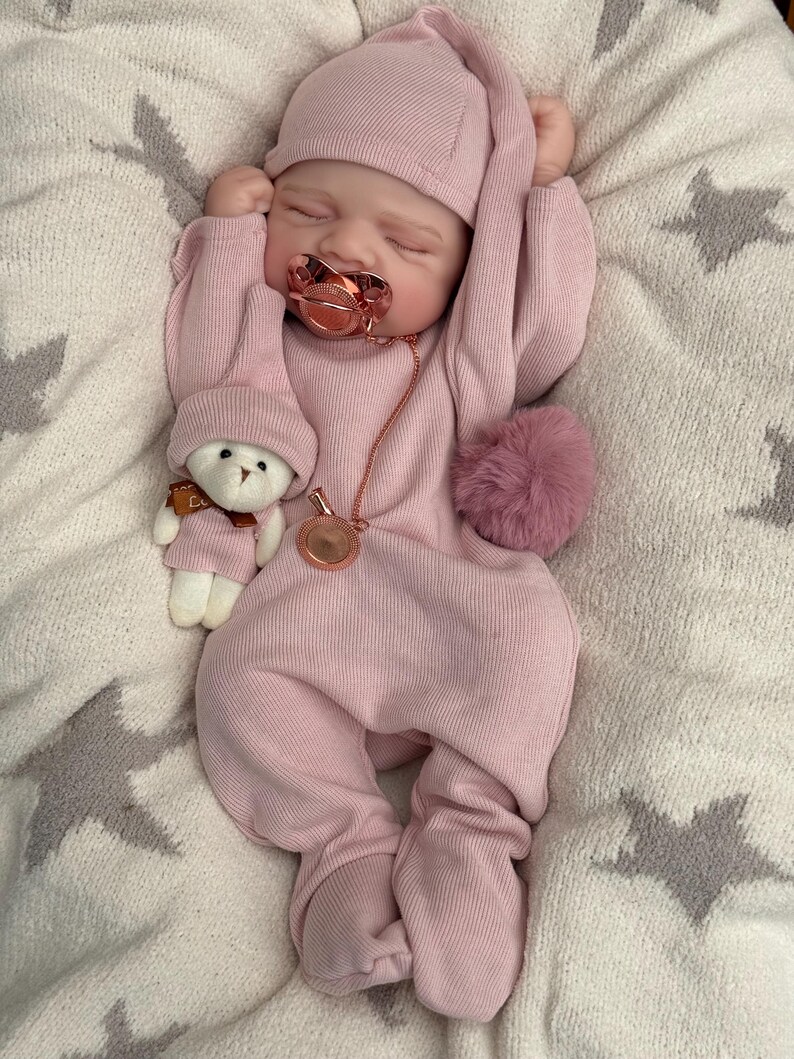 Pu&ograve; includere: Una bambola neonato addormentata che indossa una tutina a coste rosa polvere a maniche lunghe con berretto abbinato. La bambola ha un ciuccio color oro rosa con catenella e tiene in mano un piccolo orsacchiotto bianco con berretto e sciarpa.