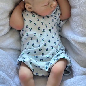 May include: A realistic baby doll with light skin and closed eyes, wearing a white short-sleeved onesie featuring a small purple and green floral pattern. The doll has a white elastic headband with a fabric flower on its head and is lying down.