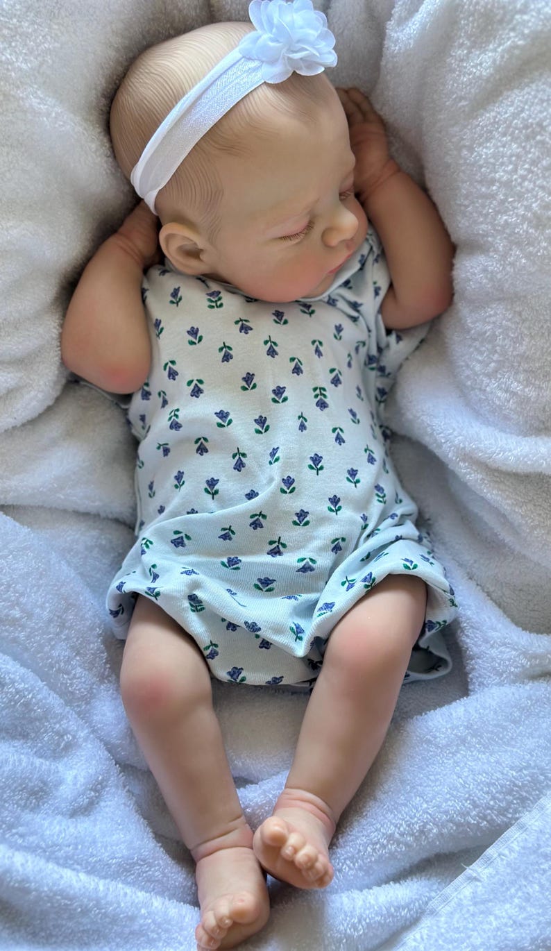May include: A realistic baby doll with light skin and closed eyes, wearing a white short-sleeved onesie featuring a small purple and green floral pattern. The doll has a white elastic headband with a fabric flower on its head and is lying down.