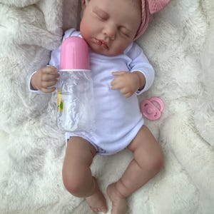 May include: A realistic baby doll with closed eyes, wearing a white long-sleeve bodysuit and a dusty pink cable-knit headband. The doll holds a clear plastic baby bottle with a pink cap and has a small pink pacifier resting nearby on the fabric.